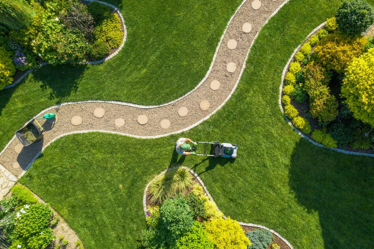Aerial view of a person mowing a landscaped garden, featuring curved stone paths, green grass, and various plants and shrubs. The garden's design includes circular stepping stones and neatly arranged greenery.