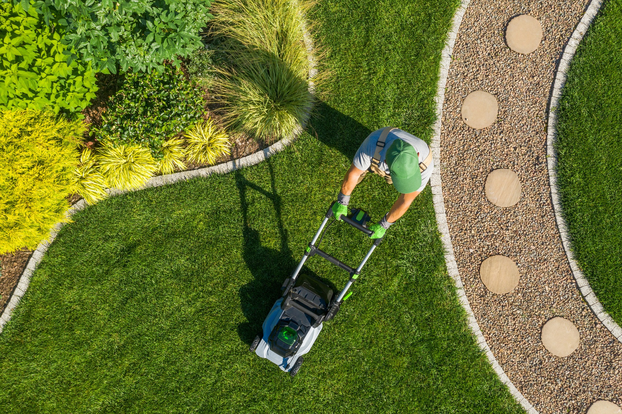 A person in a green hat and gloves is pushing a lawn mower over a neatly manicured lawn with curved edges, surrounded by various plants and a gravel path with circular stepping stones. The scene is viewed from above.