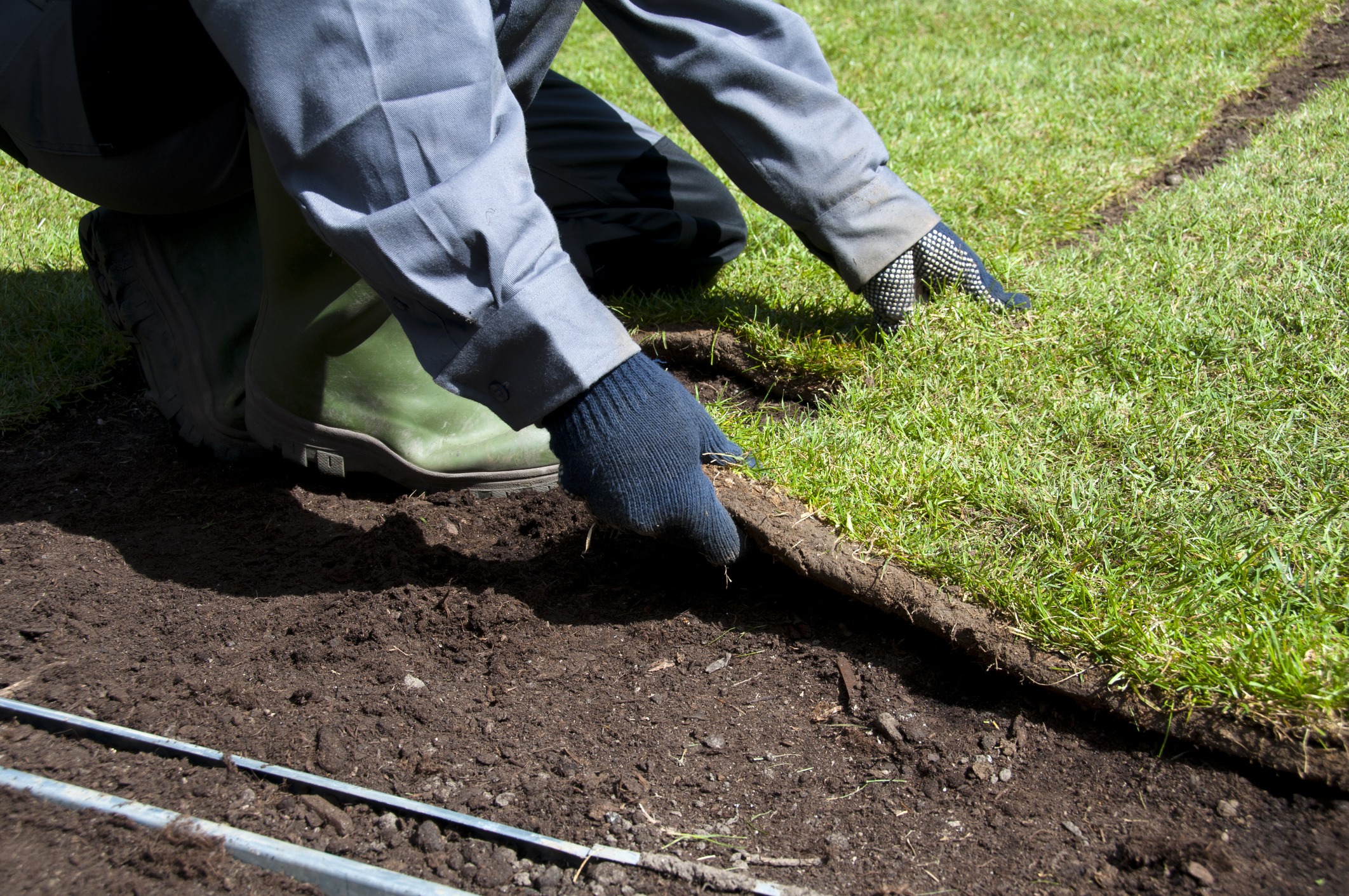 A person wearing gray clothing, gloves, and green boots is laying sod on a patch of soil. The image shows the individual adjusting a piece of turf, aligning it carefully with the ground, with visible mulch and soil lines in the foreground.