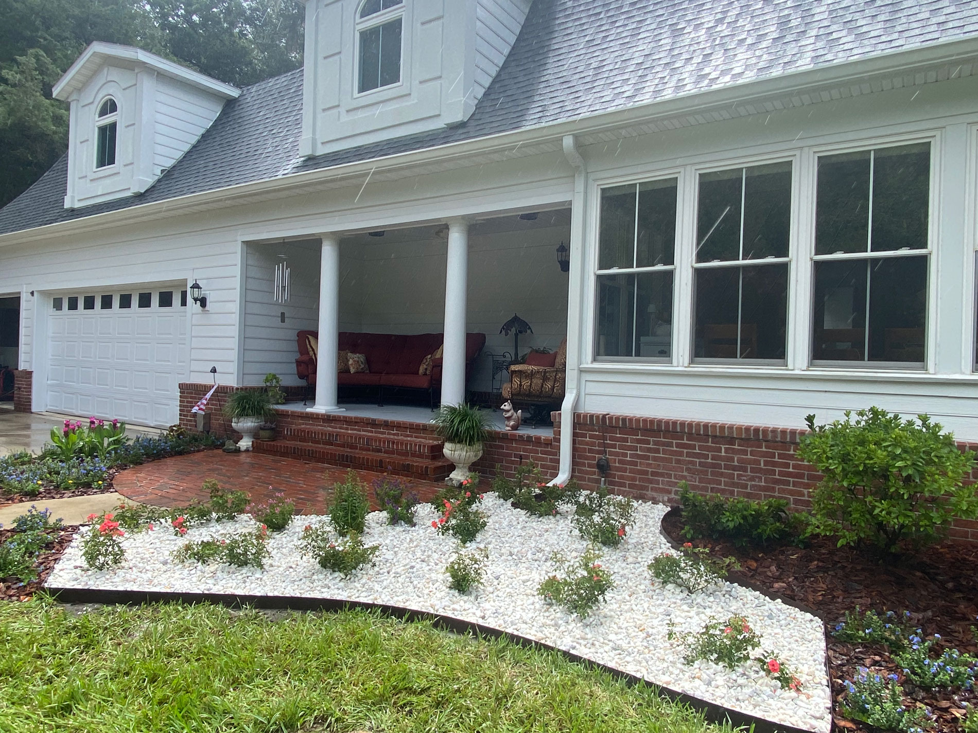 A two-story house with white siding and a brick foundation. The front porch has white columns and a red brick floor. In the garden, there are white gravel areas with small green plants and red flowers. A large garage door is on the left.